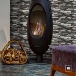 Fachaleta tipo piedra_30x45_alameda_plomeria garcia_2-ok | Plomería García Contemporary freestanding fireplace with a glass front burning logs, next to a basket of firewood and a purple upholstered stool in a living room.