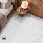 Piso ceramico color blanco-44x44-brillante-interior-porta-lamosa-ok | Plomería García Top-down view of a bedroom corner: beige bed on the left, round wooden nightstand with a lamp and small decorative items, set on a white marble tiled floor.